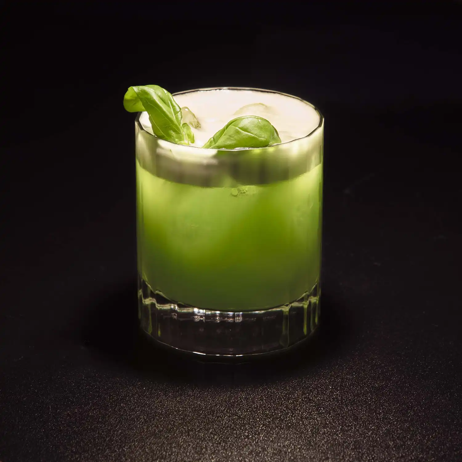 Verre à gin vert pastel, garniture de feuilles de basilic, sur fond sombre, mettant en valeur une boisson artisanale rafraîchissante.