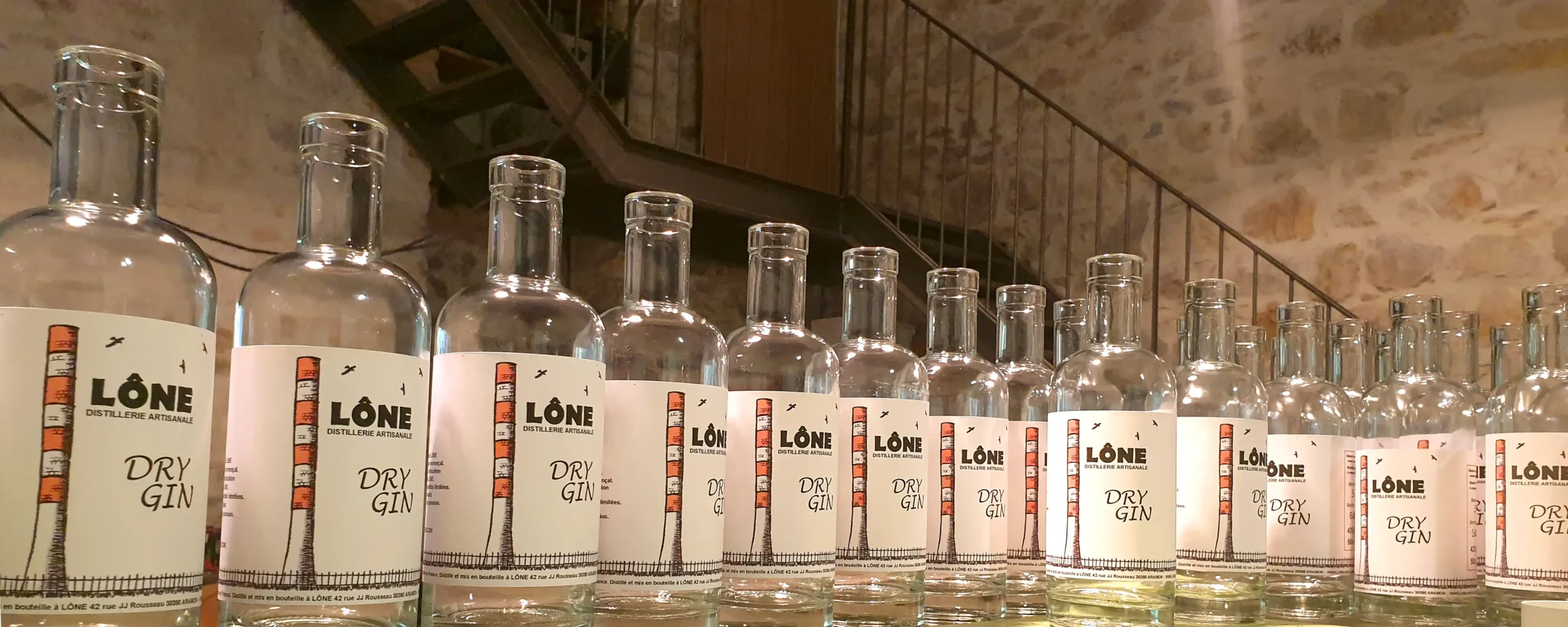Bouteilles de gin en rangée dans une cave ou bar, étiquettes lisibles indiquant « LØNE Dry Gin », ambiance chaleureuse et rustique. Gin degré alcool évoqué par les bouteilles alignées.