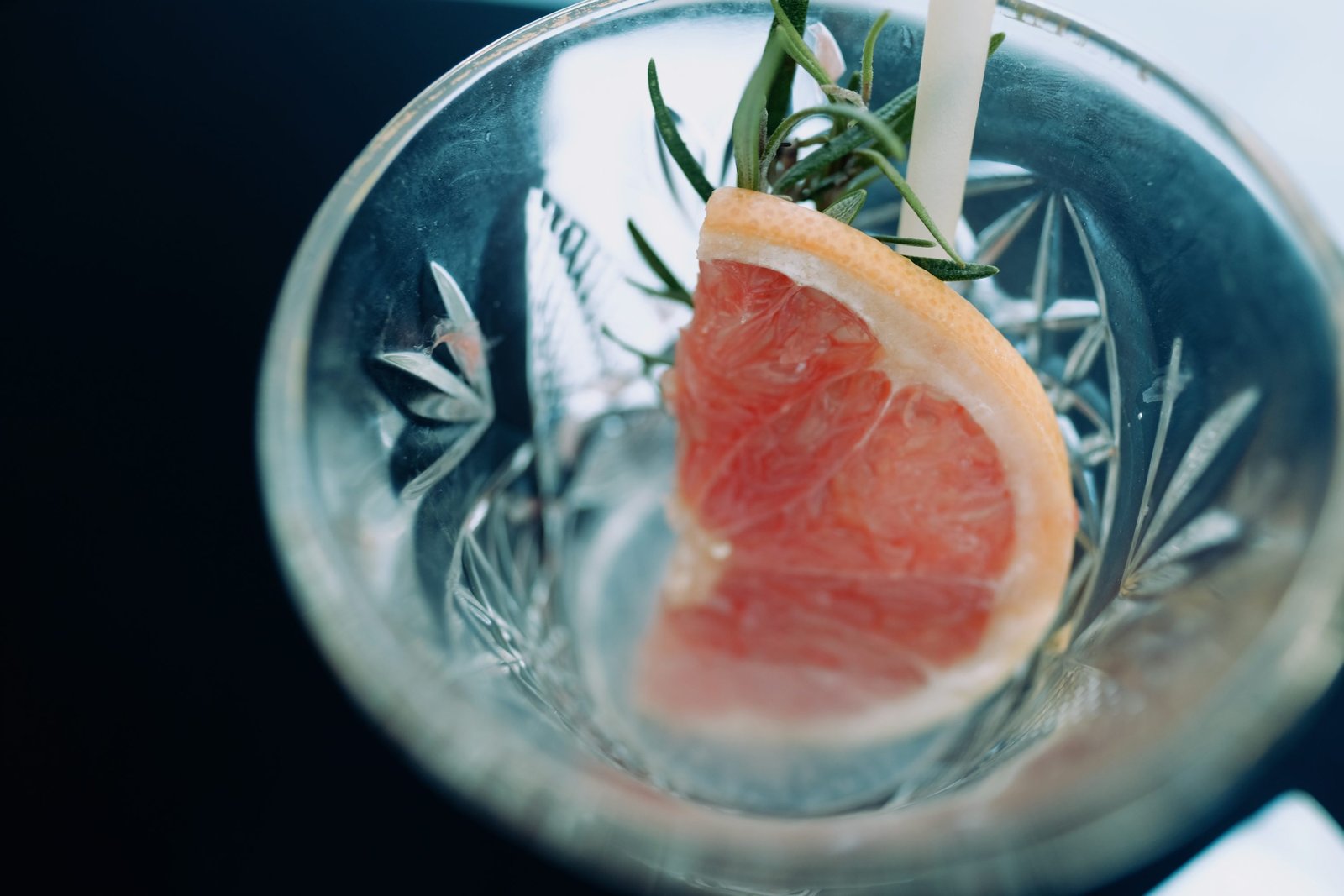 Gros plan sur un verre transparent refroidi contenant un quartier de pamplemousse rose et uneaments herbes aromatiques, avec une paille, prêt à déguster.